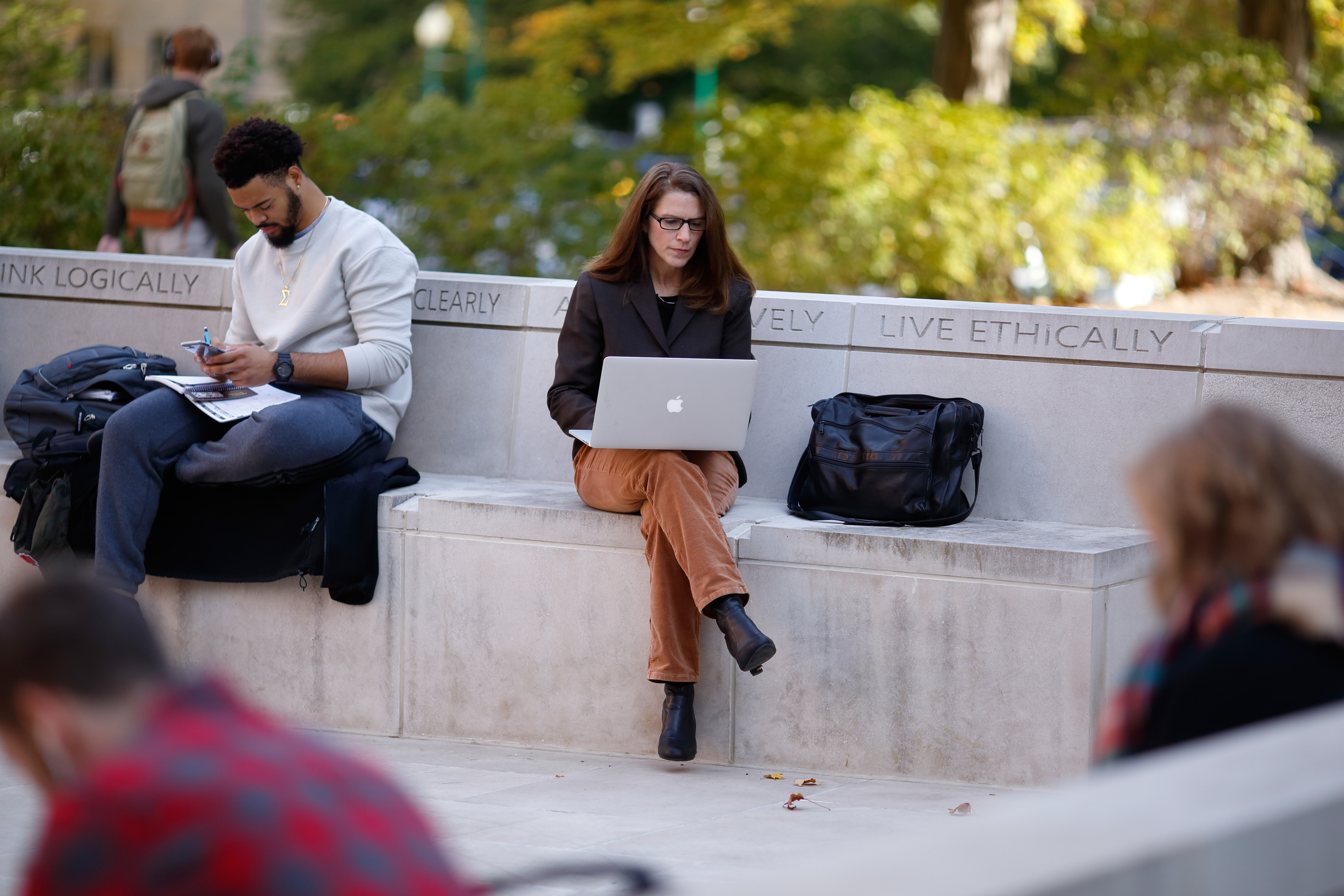 Students sitting on a bench inscribed with the College's Foundational Skills of Think Logically, Communicate Clearly, Act Creatively, and Live Ethically. Missing from the image is Question Critically.